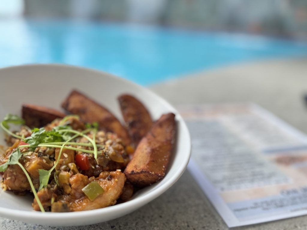 Vegetable stir-fry with plantains served in a white bowl, with a pool in the background, highlightin.